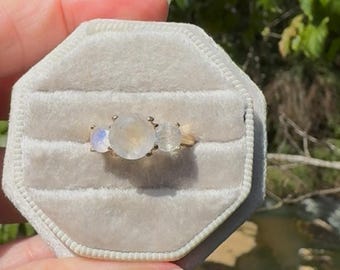 May include: A gold ring featuring three round moonstones set in a row. The center stone is the largest, flanked by two smaller moonstones on either side. The stones exhibit a translucent, iridescent quality with subtle blue and white flashes.