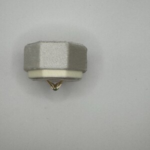 May include: A light gray, octagonal velvet ring box featuring a textured white ribbon trim around the base. A thin gold ring with a small, dark-colored stone is positioned at the bottom center, partially visible beneath the box's edge.