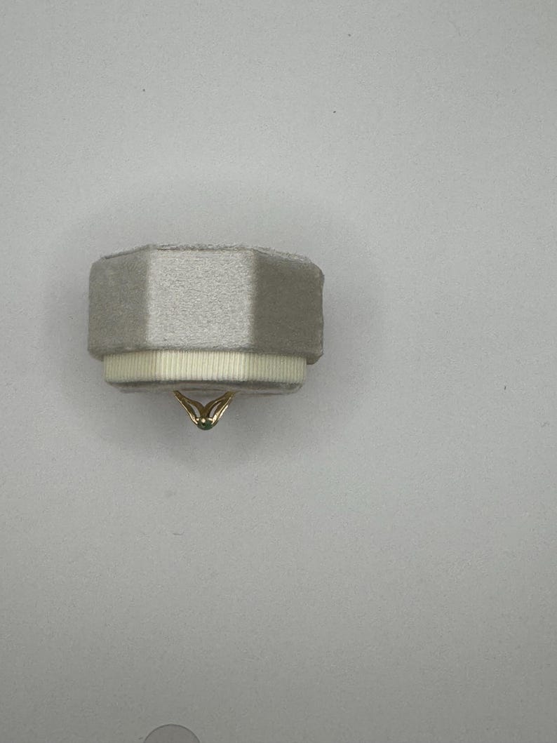 May include: A light gray, octagonal velvet ring box featuring a textured white ribbon trim around the base. A thin gold ring with a small, dark-colored stone is positioned at the bottom center, partially visible beneath the box's edge.