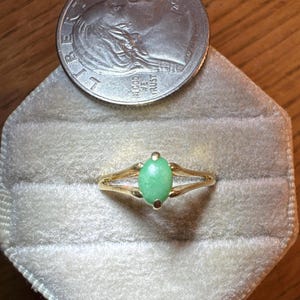 May include: A gold ring featuring a small, oval-shaped, light green stone set in a four-prong mounting. The ring is positioned below a 2022 United States dime, which provides a sense of scale for the jewelry piece against the velvet surface.