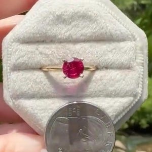 May include: A gold ring with a vibrant red gemstone, presented in a gray velvet ring box. The ring's band is thin and gold, and the gemstone is round. A US quarter is in the foreground.