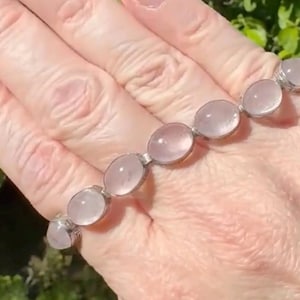 May include: A silver bracelet featuring oval, pale pink gemstones. The gemstones are set in individual silver settings and connected by small silver links. The bracelet is displayed on a hand, with a natural green background.