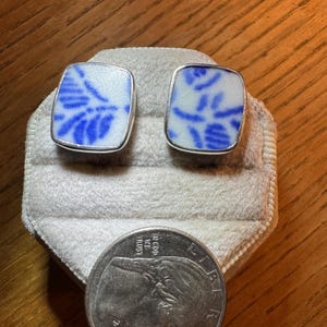 May include: Two rectangular stud earrings featuring white ceramic with blue floral patterns set in silver-toned metal frames. A 2022 United States quarter coin is placed below the earrings for scale, showing the relative size of the jewelry pieces.