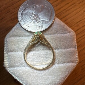 May include: A gold ring featuring a small, round, light green gemstone set in a prong setting. The ring is positioned next to a United States quarter coin, which serves as a scale reference for the size of the jewelry piece.