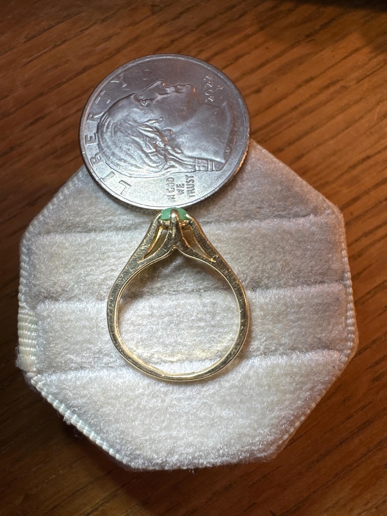 May include: A gold ring featuring a small, round, light green gemstone set in a prong setting. The ring is positioned next to a United States quarter coin, which serves as a scale reference for the size of the jewelry piece.