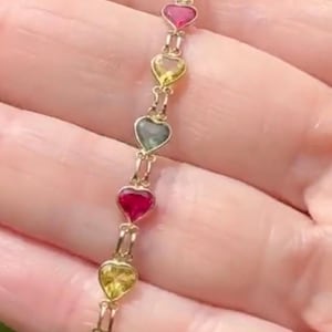 May include: A gold chain bracelet featuring a series of heart-shaped gemstones in alternating colors of red, yellow, and green. Each heart is linked by small gold metal loops, creating a delicate and continuous strand of colorful heart charms.