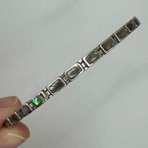 May include: Silver-toned bracelet featuring rectangular abalone shell inlays. The bracelet has a link design with a repeating pattern of the iridescent shell pieces. The bracelet is held against a light background.