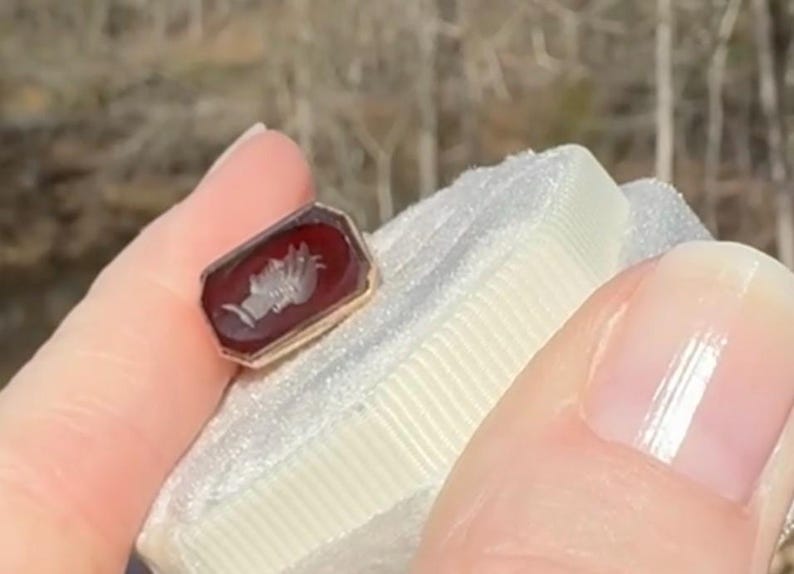 Pu&ograve; includere: Un anello rettangolare dorato con una pietra rossa. La pietra presenta un'incisione bianca dettagliata di una mano che tiene una torcia. L'anello &egrave; tenuto contro una superficie bianca strutturata.