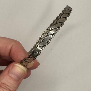 May include: A silver-toned bracelet with a braided chain design. The bracelet is made of interlocking oval links, creating a textured surface. The jewelry is shown against a neutral background.