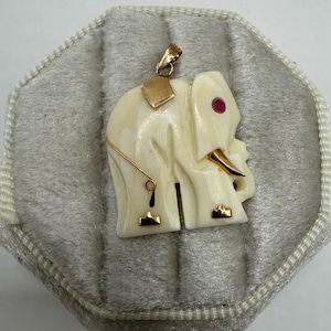 May include: Ivory-colored elephant pendant with gold accents, including a gold bale, ear, and decorative elements. The elephant has a red gemstone eye. The pendant is displayed on a gray, textured surface.