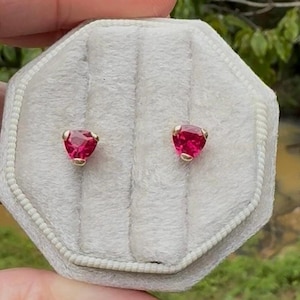 May include: A pair of heart-shaped, ruby-red gemstone stud earrings. The gemstones are set in gold-colored metal. The earrings are displayed in a gray, velvet-lined jewelry box.