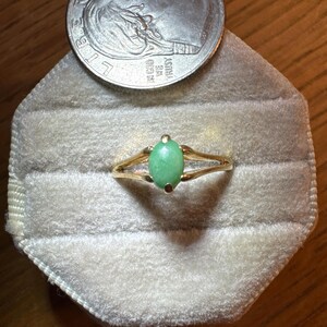 May include: A gold ring featuring a single oval-shaped light green stone set in a four-prong setting. The ring is positioned below a 2022 United States dime, which provides a scale reference for the size of the jewelry piece.