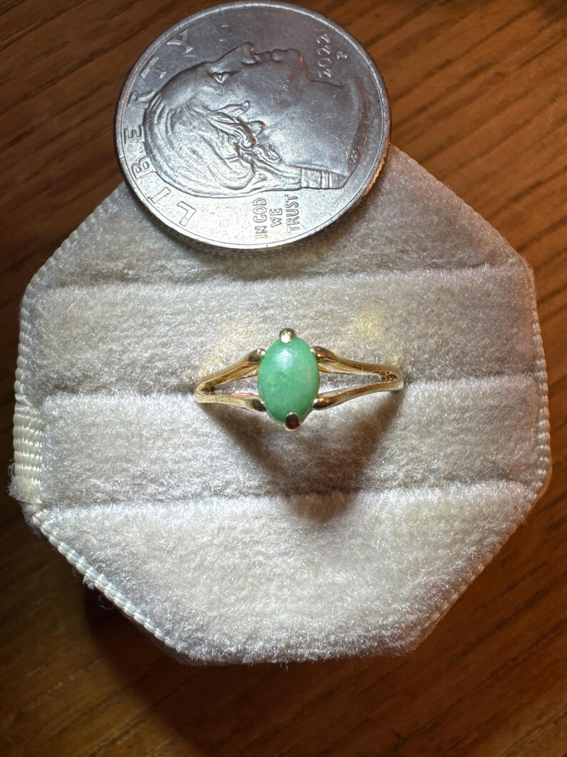 May include: A gold ring featuring a single oval-shaped light green stone set in a four-prong setting. The ring is positioned below a 2022 United States dime, which provides a scale reference for the size of the jewelry piece.