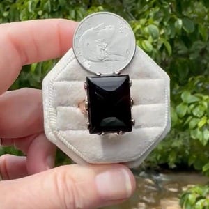 May include: A ring featuring a large, rectangular, faceted black gemstone set in a metal band. The ring is displayed on a white velvet ring holder next to a United States nickel coin for scale, which has a diameter of approximately 0.835 inches.