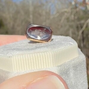 Pu&ograve; includere: Un anello rettangolare con una fascia dorata e una pietra trasparente incisa. La pietra ha una tonalit&agrave; rossa e presenta un'incisione dettagliata. L'anello &egrave; appoggiato su una superficie bianca strutturata.