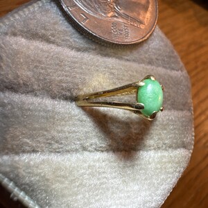 May include: A gold-toned ring featuring a smooth, oval-shaped light green cabochon stone set in a four-prong setting with a split shank band, positioned next to a United States quarter coin for scale.