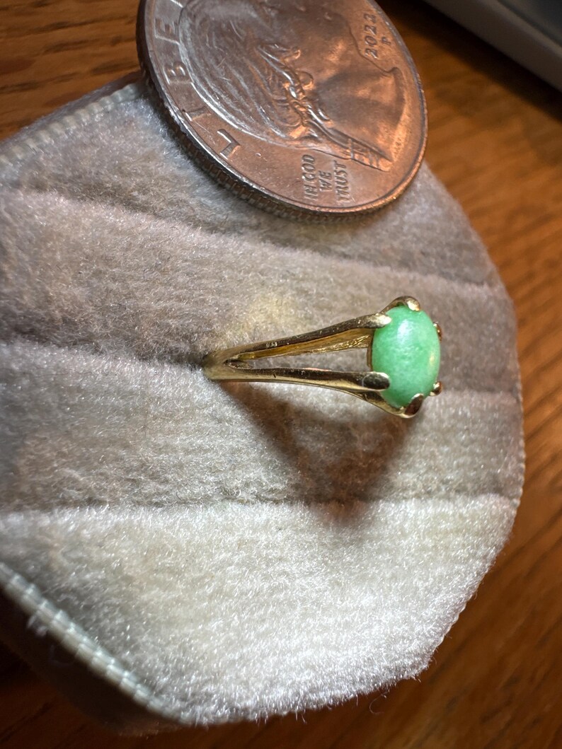 May include: A gold-toned ring featuring a smooth, oval-shaped light green cabochon stone set in a four-prong setting with a split shank band, positioned next to a United States quarter coin for scale.