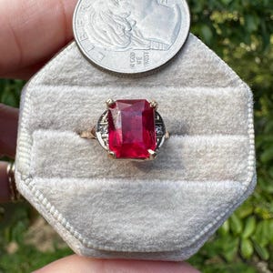May include: A gold ring featuring a large, rectangular, faceted red gemstone set in the center, flanked by two small, clear, round accent stones on each side. The ring is displayed on a beige velvet ring holder next to a 2022 U.S. dime coin.