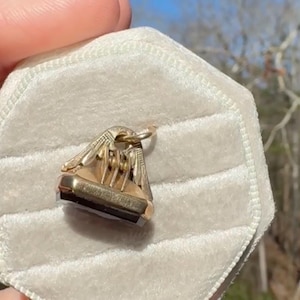 Pu&ograve; includere: Un ciondolo dorato con una pietra rettangolare scura &egrave; esposto su una superficie di velluto bianco. Il ciondolo presenta un design ornato e un anello per una catena. Lo sfondo &egrave; sfocato.