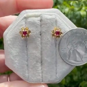 May include: A pair of gold-colored flower-shaped stud earrings with a small, round, red gemstone in the center of each flower. The earrings are displayed on a gray velvet surface next to a US quarter coin.