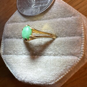 May include: A gold ring featuring a smooth, oval-shaped light green stone set in a four-prong mounting. The band has a split design on the sides. A United States quarter coin is positioned directly above the ring for a size comparison.