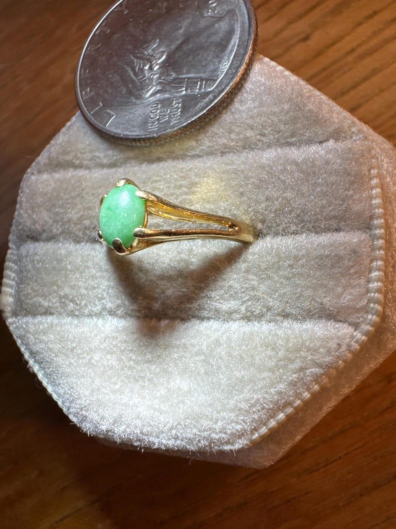May include: A gold ring featuring a smooth, oval-shaped light green stone set in a four-prong mounting. The band has a split design on the sides. A United States quarter coin is positioned directly above the ring for a size comparison.