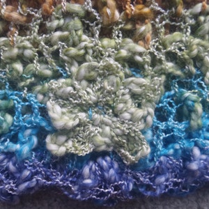 May include: Close-up of a hand-knitted textile with a flower design. The yarn is a mix of blues, greens, and browns, creating a textured, multi-colored effect. The flower is a prominent, three-dimensional element.