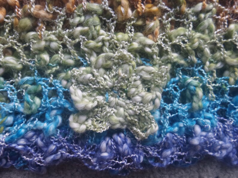 May include: Close-up of a hand-knitted textile with a flower design. The yarn is a mix of blues, greens, and browns, creating a textured, multi-colored effect. The flower is a prominent, three-dimensional element.