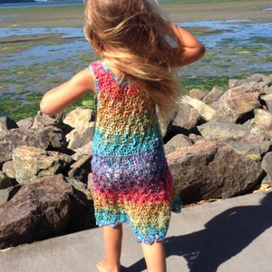 May include: A sleeveless, rainbow-colored crochet dress. The dress features a vibrant spectrum of colors, including red, orange, yellow, green, blue, and purple. The dress is worn by a person standing on a concrete surface near a body of water.