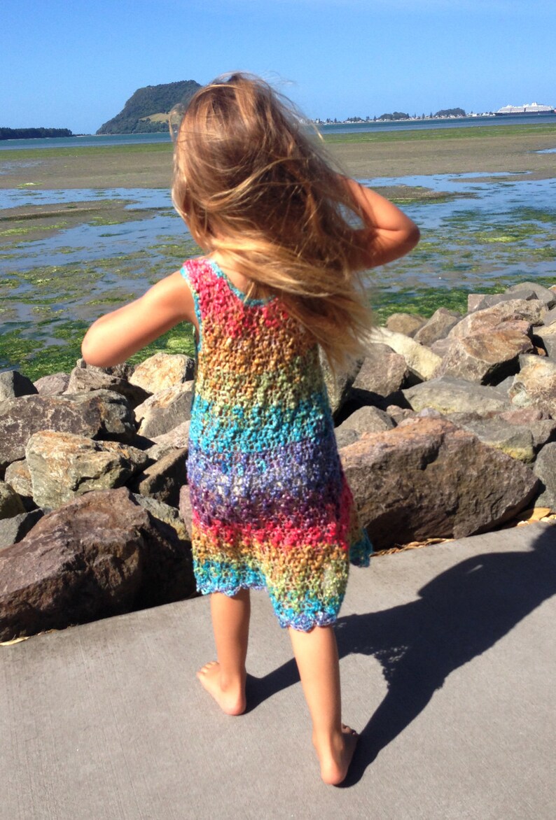 May include: A sleeveless, rainbow-colored crochet dress. The dress features a vibrant spectrum of colors, including red, orange, yellow, green, blue, and purple. The dress is worn by a person standing on a concrete surface near a body of water.