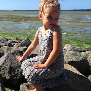 May include: A young girl wearing a gray crochet dress with a flower design sits on rocks by the water. The dress has a ruffled hem and a sleeveless design. The girl has her hair pulled back in a bun.
