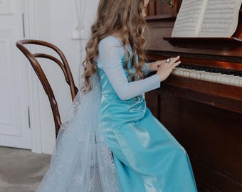 Könnte beinhalten: Ein hellblaues Satinkleid mit langen, transparenten Ärmeln und einem passenden, durchsichtigen Umhang mit weißen Schneeflockenmustern. Das Kleid hat ein eng anliegendes Oberteil und einen bodenlangen Rock, kombiniert mit hellen flachen Schuhen.