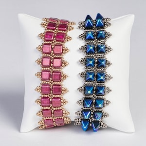 May include: Two beaded bracelets, one in shades of pink and gold, the other in blue and silver, rest on a white pillow. The pink bracelet features square beads, while the blue bracelet has square and triangular beads.