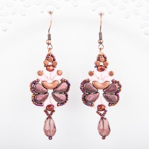 May include: A pair of dangle earrings with a pink, copper, and purple color scheme. The earrings feature a teardrop-shaped crystal and a beaded design that resembles a flower.