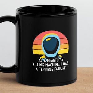 May include: Black coffee mug with a retro sunset design and a cartoon astronaut. The text reads "As a heartless killing machine, I was a terrible failure."