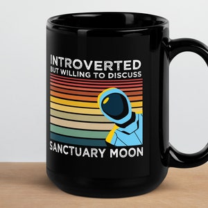 May include: Black coffee mug with a retro design featuring an astronaut and the text "Introverted but willing to discuss Sanctuary Moon".