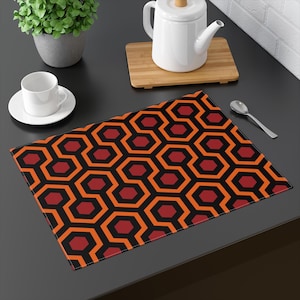 May include: A rectangular placemat featuring a repeating geometric pattern of interlocking hexagons in black, orange, and dark red. The design consists of thick black outlines forming a continuous maze-like structure with orange and red centers.