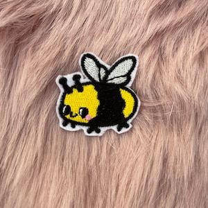 May include: A yellow and black embroidered bee patch with a cute cartoon face. The bee has white wings and is on a fuzzy pink background.