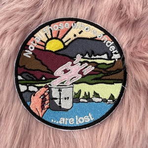 May include: Embroidered patch featuring a colourful landscape scene, a mug of steaming liquid, and the text "Not all those who wander... are lost".