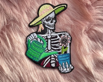 May include: An embroidered patch featuring a white skeleton wearing a yellow sun hat with a green brim. The skeleton holds a bright green watering can in one hand and a blue flower pot containing a pink flower in the other hand.