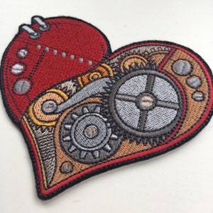 Könnte beinhalten: Ein herzförmiger Stickaufnäher mit mechanischem Design. Die linke Seite ist rot mit silbernen Nietendetails, während die rechte Seite komplizierte Zahnrad- und Getriebemotive in Bronze und Silber zeigt, die durchgehend mit schwarzem Garn umrandet sind.