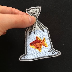 May include: An embroidered patch depicting a goldfish in a plastic bag. The goldfish is orange and yellow with black eyes. The bag is white with a grey outline.