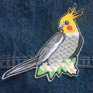 May include: Embroidered iron-on patch of a grey and yellow cockatiel perched on a branch with white flowers. The bird has a yellow head and a grey body with white feathers.