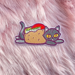 May include: A gray cat embroidered patch with yellow eyes. The cat is curled up inside a brown taco with red tomato, green lettuce, and white cheese fillings.
