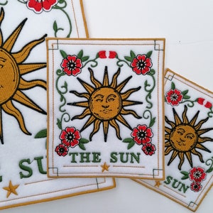 May include: Three rectangular embroidered patches on a white background, each featuring a central golden sun with a human face, surrounded by red flowers and green vines. The text THE SUN is embroidered in green at the bottom of each patch.