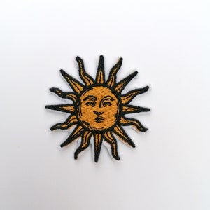 Op de afbeelding: Een geborduurde gele zon met een gezicht en zwarte stralen. De zon is op een witte achtergrond.