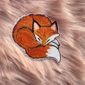May include: Embroidered patch of a curled up orange fox with white accents, sleeping on a pink fluffy surface.
