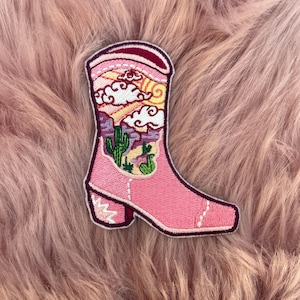 May include: Pink embroidered iron-on patch of a cowboy boot with a desert landscape design. The boot has a pointed toe and a heel. The landscape features a sunset, clouds, and cacti.