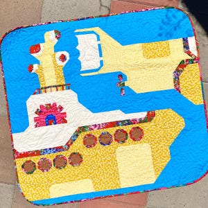 May include: A yellow and white quilted throw blanket with a blue background featuring a submarine design. The submarine has a red and pink periscope and is surrounded by yellow and white flowers.