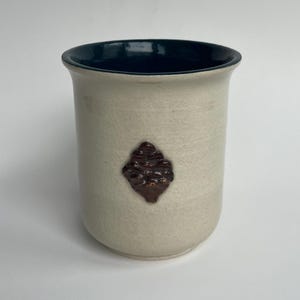 May include: A ceramic tumbler with a cream-coloured exterior and a dark blue interior. A brown, textured, diamond-shaped design is centered on the tumbler's exterior. The tumbler is a cylindrical shape.